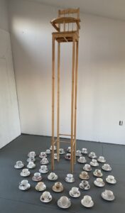 Image of a mixed media and found object piece of artwork by Frances Key. Includes a high chair with long legs set in the middle of teacups and saucers spread out on the floor