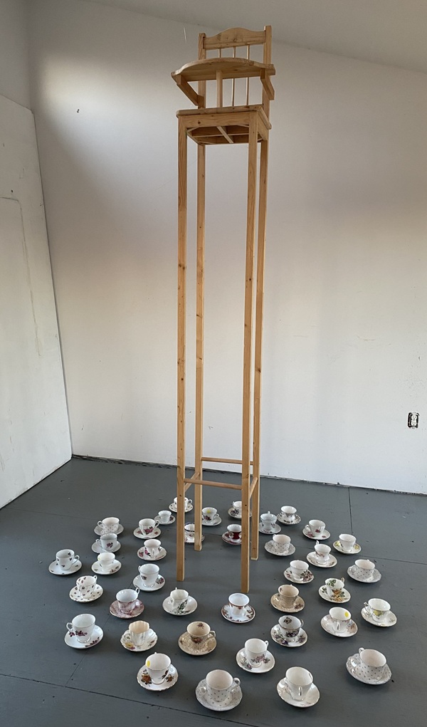 Image of a mixed media and found object piece of artwork by Frances Key. Includes a high chair with long legs set in the middle of teacups and saucers spread out on the floor
